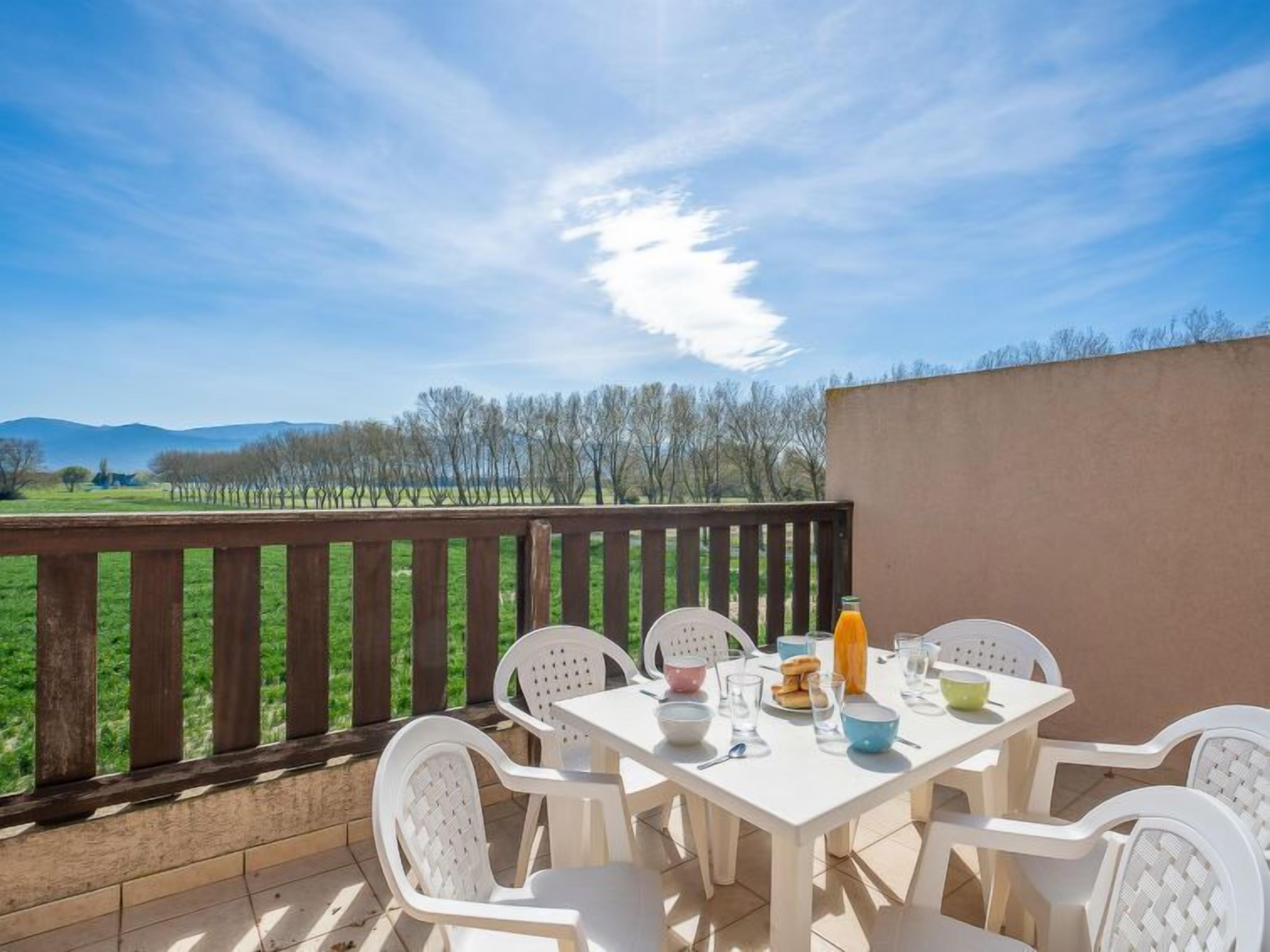 Een terras met een ontbijttafel, omringd door witte stoelen, met uitzicht op een groon landschap met bomen en bergen in de verte.