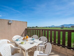 Pátio com mesa branca posta para o café da manhã, rodeada por cadeiras brancas, com vista para um campo verde e montanhas.