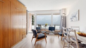 Een moderne woonkamer met houten vloer, een eethoek met witte stoelen, een televisie en een groot raam met uitzicht op bergen.
