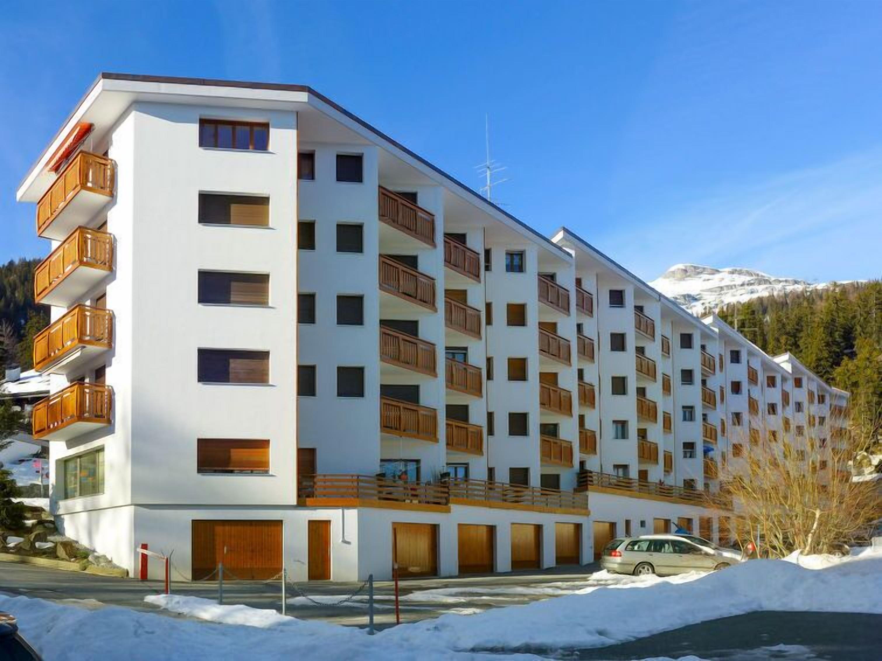 Un hôtel à plusieurs étages doté de balcons, avec un télésiège visible au loin et un paysage enneigé.