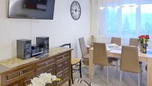 A modern living room with a flat-screen TV, a clock, a dining area with a table and chairs, and a flower vase. - Barzettes-Vacances 553 / Entrée N° 14 by Interhome (Crans-Montana)