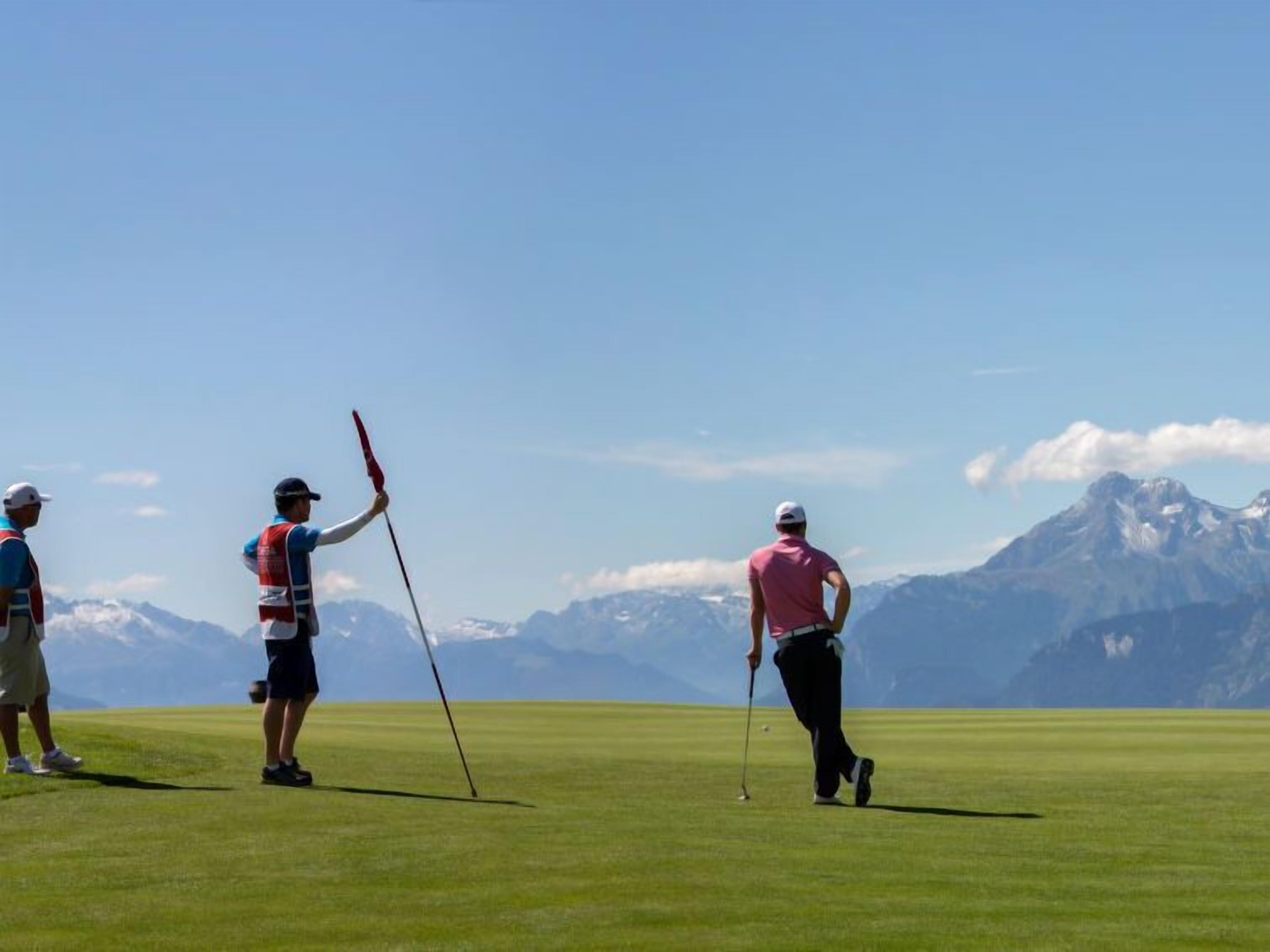 Un terrain de golf où l’on voit des joueurs en action, un ciel dégagé et des montagnes en arrière-plan.