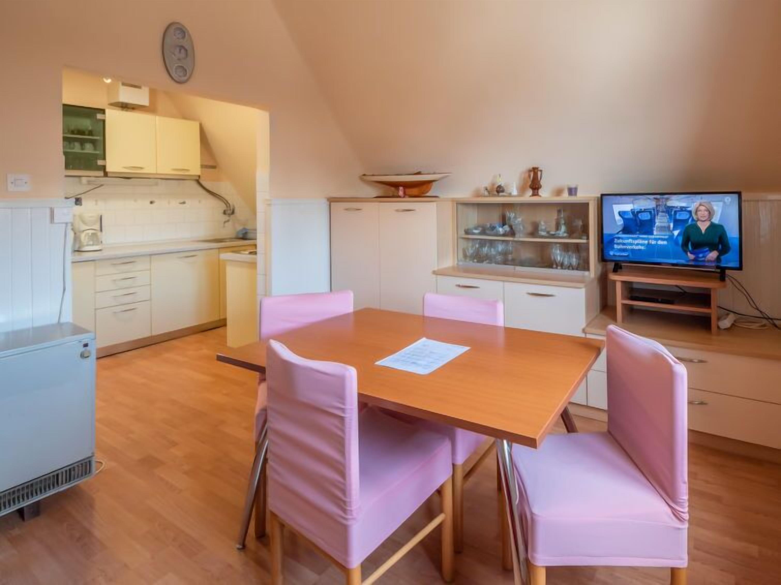 A compact kitchen with white cabinets, a small refrigerator, and a dining area with a wooden table and pink chairs.