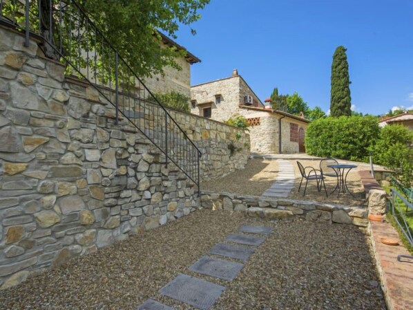 A stone staircase leading to a building with a rustic exterior, a paved patio with outdoor seating, and a tall tree in the background.