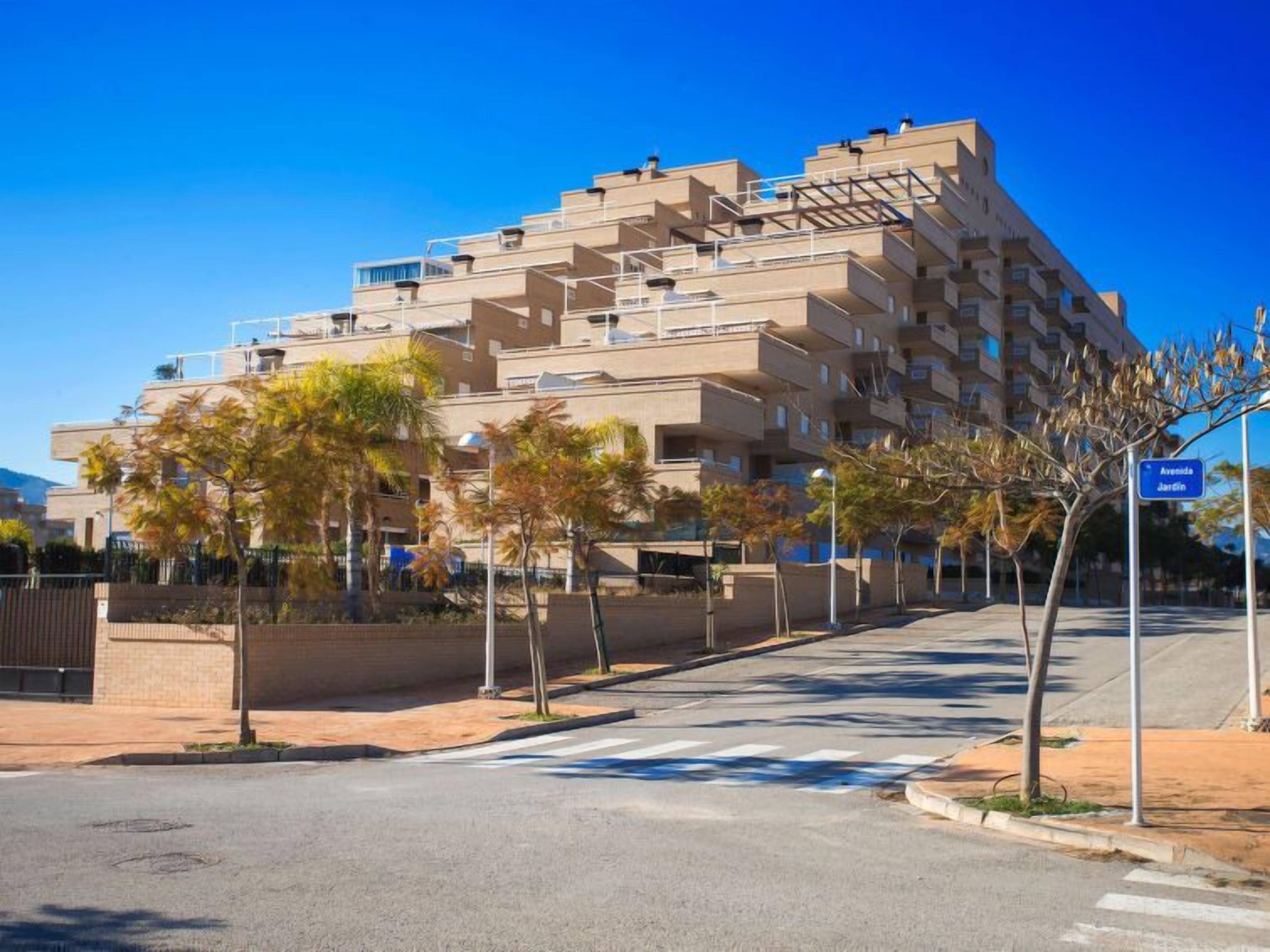 Un edificio residencial de varios pisos con balcones, cielo despejado, calle con árboles y acera.
