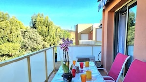 Un balcón con una mesa dispuesta para dos personas, con un mantel decorado con motivos frutales, una orquídea en maceta y varios vasos.