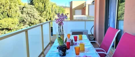 A balcony with a table set for two, featuring a tablecloth with fruit designs, a potted orchid, and various drinkware.
