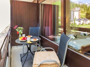 A balcony with a table set for two, a cushioned chair, and a view of a building and trees.