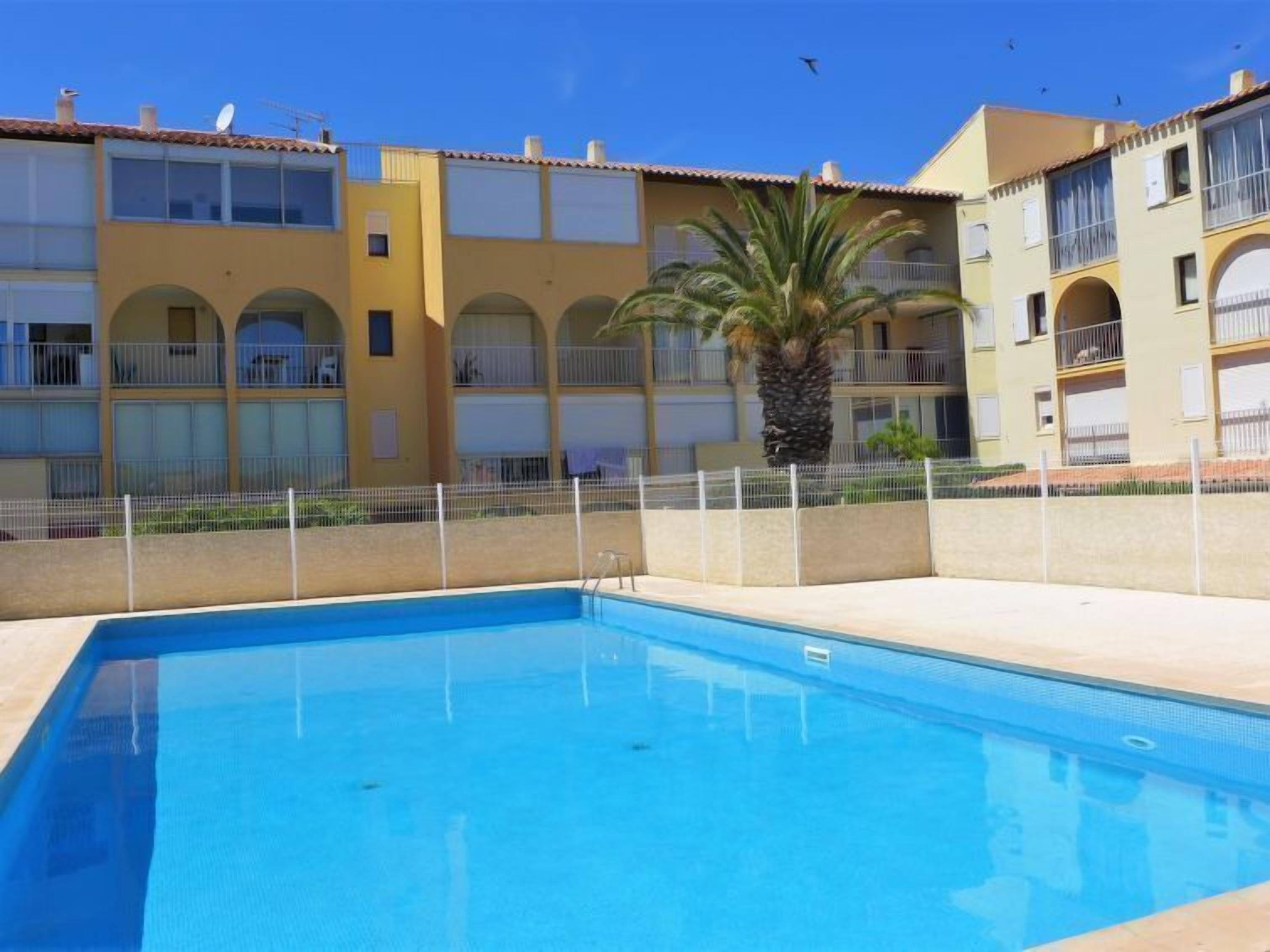 A swimming pool with a clear blue water surface, surrounded by a white fence and a building with balconies and arched windows.