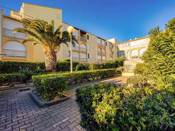 A courtyard with neatly trimmed hedges, a palm tree, and a paved walkway. - Les Maisons sur la Plage by Interhome (Narbonne-Plage)