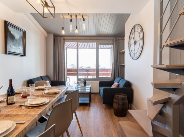 A modern living room with a dining area, a sofa, a clock, and a view of the city through large windows.