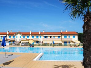 Zona de piscina con terraza de madera, una palmera y una hilera de edificios con tejados rojos y ventanas azules.