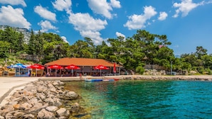 A coastal area with clear turquoise water, a rocky shoreline, and a red-roofed building with outdoor seating. - Apartment Delfin by Interhome (Karlobag)