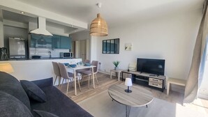 Ein modernes Wohnzimmer mit einem dunklen Sofa, einem runden Couchtisch und einem Fernseher. Es gibt einen Essbereich mit Tisch und Stühlen sowie eine Küche mit Mikrowelle und Schränken.