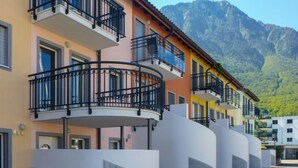 A row of multi-story buildings with balconies, a table with chairs, and a mountain backdrop.