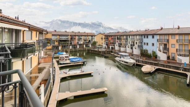 A canal with boats docked, surrounded by multi-story buildings with balconies, and mountains in the background.