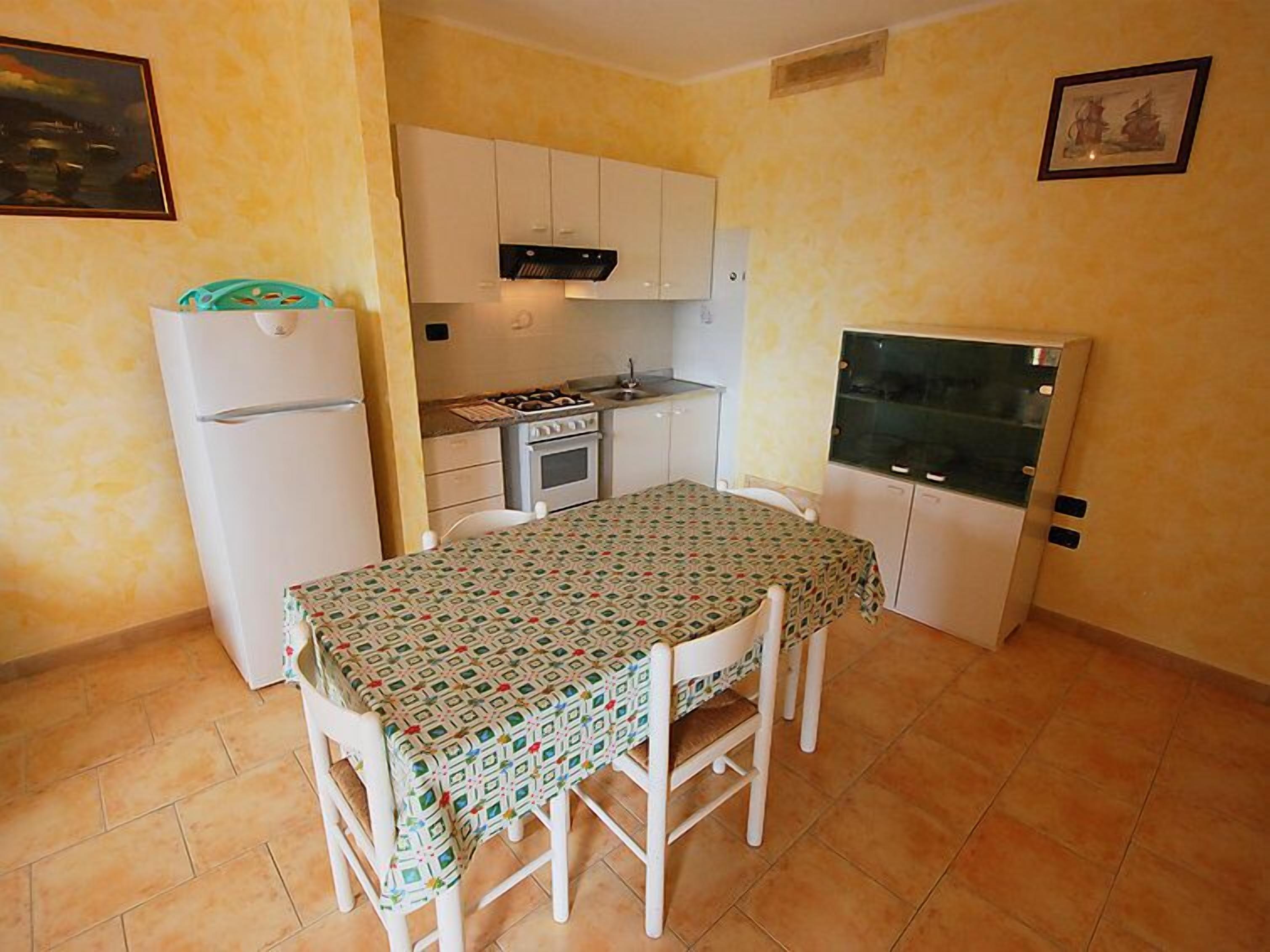 Een kleine keuken met witte kasten, een fornuis en een koelkast. Er is een eettafel met stoelen en een tafelkleed met een patroon.