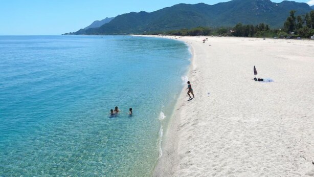 En strand med kristallklart turkost vatten, en sandstrand och berg i bakgrunden.