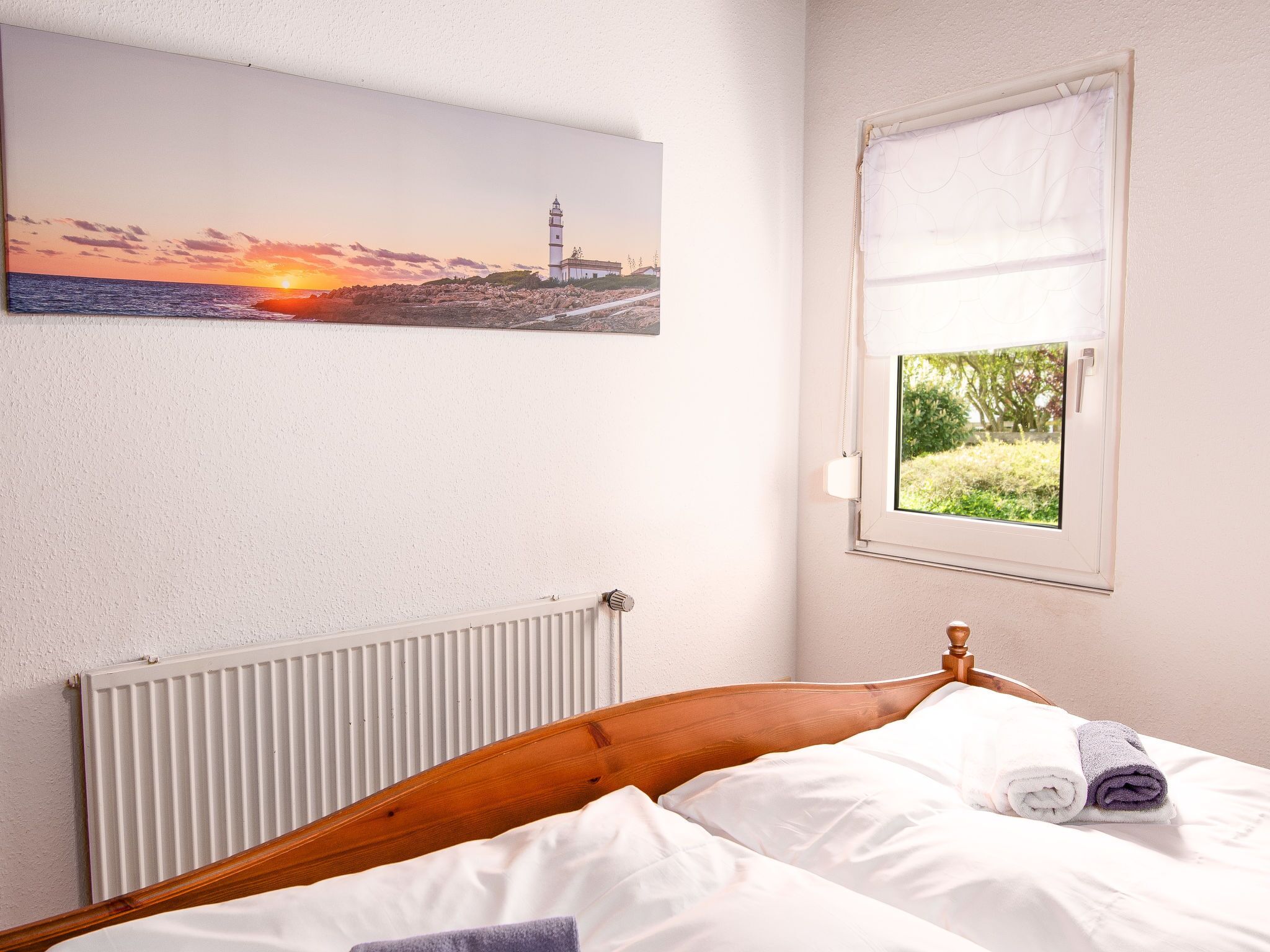 Ein Schlafzimmer mit einem hölzernen Bett, weißen Leinen, einem Heizkörper, einem Fenster mit Blick auf Grünflächen und einem Sonnenuntergangsgemälde an der Wand.