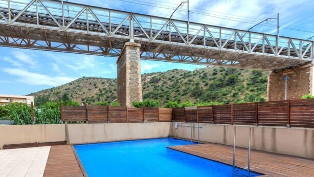 A swimming pool with a wooden deck, a stone pillar, and a metal bridge overhead. - Escorxador by Interhome (Colera)