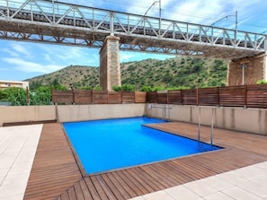 A swimming pool with a wooden deck, a stone pillar, and a metal bridge overhead.