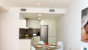 A compact kitchen with modern appliances, a dining area with a glass table and white chairs, and a small seating area with a wooden table and chairs. - Escorxador by Interhome (Colera)