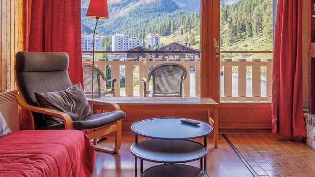 A cozy room with a wooden interior, a red chair, a small round table, and a view of mountains and buildings through large windows. - Dents Rousses H2 by Interhome (Siviez-Nendaz)
