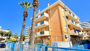 Un immeuble à plusieurs étages, doté de balcons, de palmiers et surplombant un ciel d’un bleu limpide.