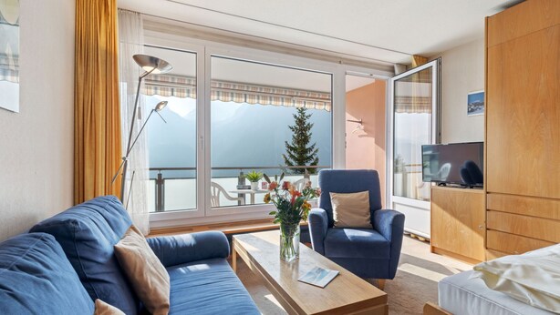 A modern hotel room with a sofa, armchair, wooden table, and a TV on a wooden cabinet. - Promenade Apt 306 by Interhome (Arosa)