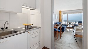 A modern kitchen with white cabinets, a stainless steel sink, and an oven. A dining area with a blue sofa and a table is visible through an open door. - Promenade Apt 306 by Interhome (Arosa)