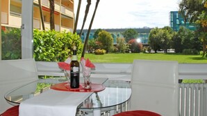 A glass table with a bottle and glasses, white chairs, and a view of a garden and buildings. - Residenza Lido Appartment 009 by Interhome (Locarno)