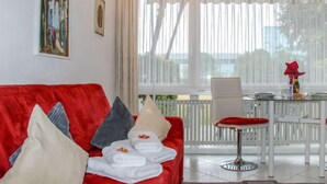 A red sofa with pillows, a dining table with chairs, and a large window with curtains. - Residenza Lido Appartment 009 by Interhome (Locarno)