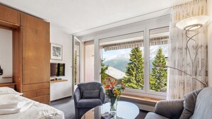 Moderni hotellihuone, jossa on sänky, sohva, tuoli, lasipöytä ja suuret ikkunat, joista avautuu näkymä vuorille.