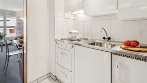 Moderni keittiö, jossa on valkoiset kaapit, ruostumattomasta teräksestä valmistettu pesuallas ja liesi. Työtasolla on tomaatteja ja leikkuulauta.