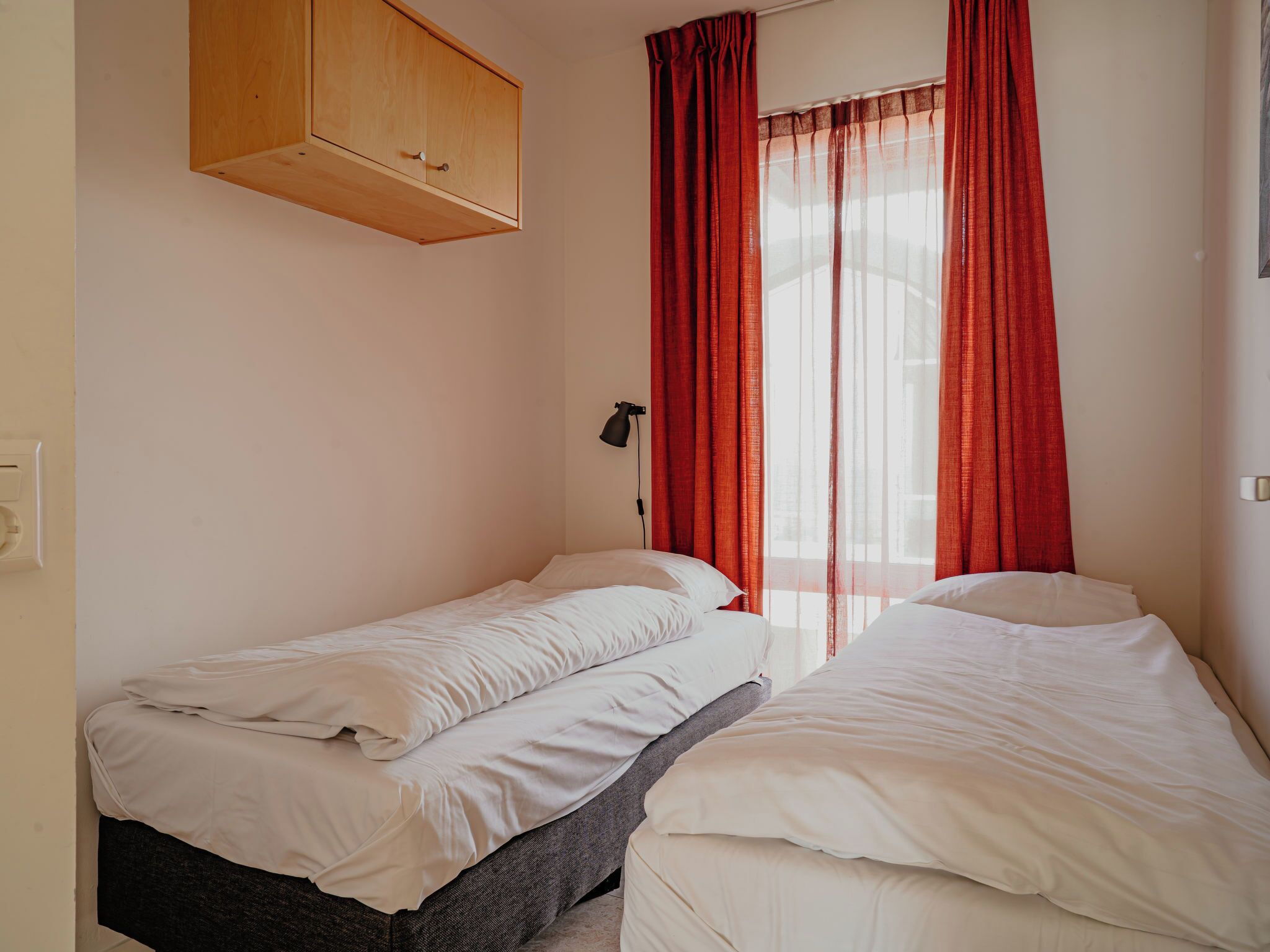 Une chambre avec deux lits simples, une armoire en bois au-dessus du lit situé à gauche, et une fenêtre avec des rideaux rouges.