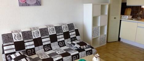 Una sala de estar con un sofá a cuadros, una mesita con un florero y una jofaina, una zona de cocina con armarios y un fregadero, y una puerta que da a otra habitación.
