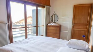 A hotel room with a large bed, a wooden dresser, a mirror, a wardrobe, and a view of buildings through a sliding glass door. - Villars Soleil B15 by Interhome (Villars)