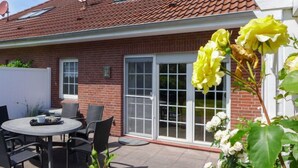 Una casa de ladrillo con techo de tejas, un patio con mesa y sillas redondas, y un jardín con flores amarillas y blancas.