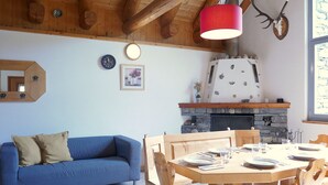 Una sala de estar acogedora con un sofá azul, una mesa de comedor de madera y sillas. Se observa una chimenea de piedra con repisa de madera.