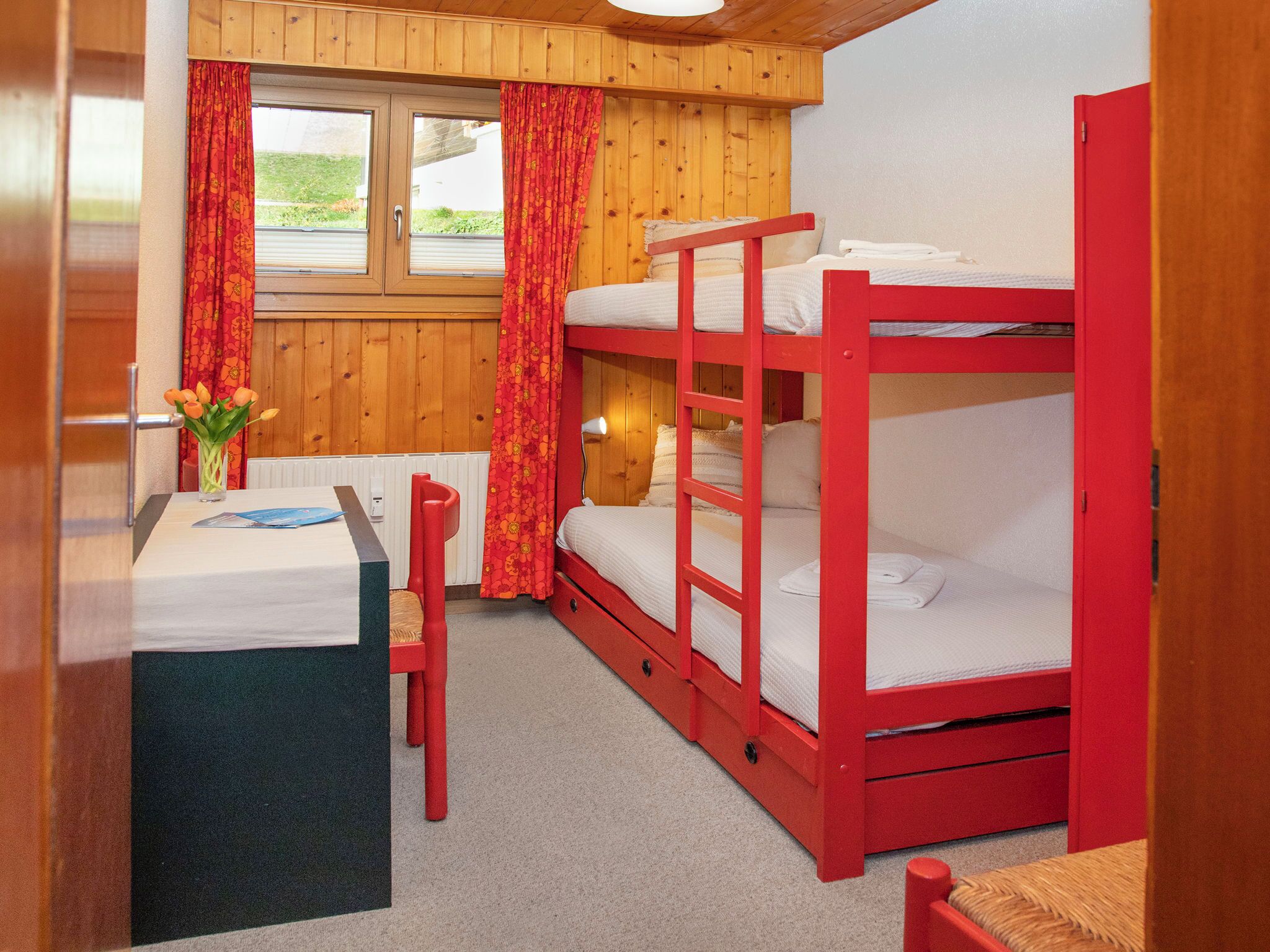 A bunk bed room with a wooden interior, a desk with a chair, and a window with curtains.