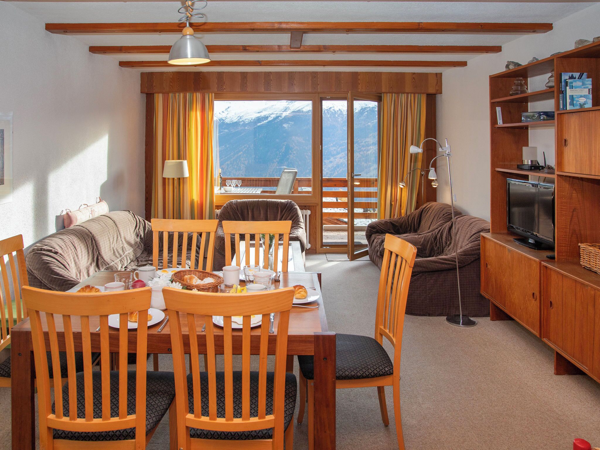 Una sala de estar acogedora con comedor, muebles de madera, televisor y vistas a montañas a través de amplias ventanas.