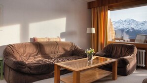 A cozy living room with a brown sofa, a wooden coffee table, and a view of snow-capped mountains through a window with curtains.