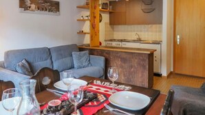 Een moderne woonkamer met een grijze bank, een houten eettafel gedekt voor twee personen, en een keuken met houten kasten en een fornuis.