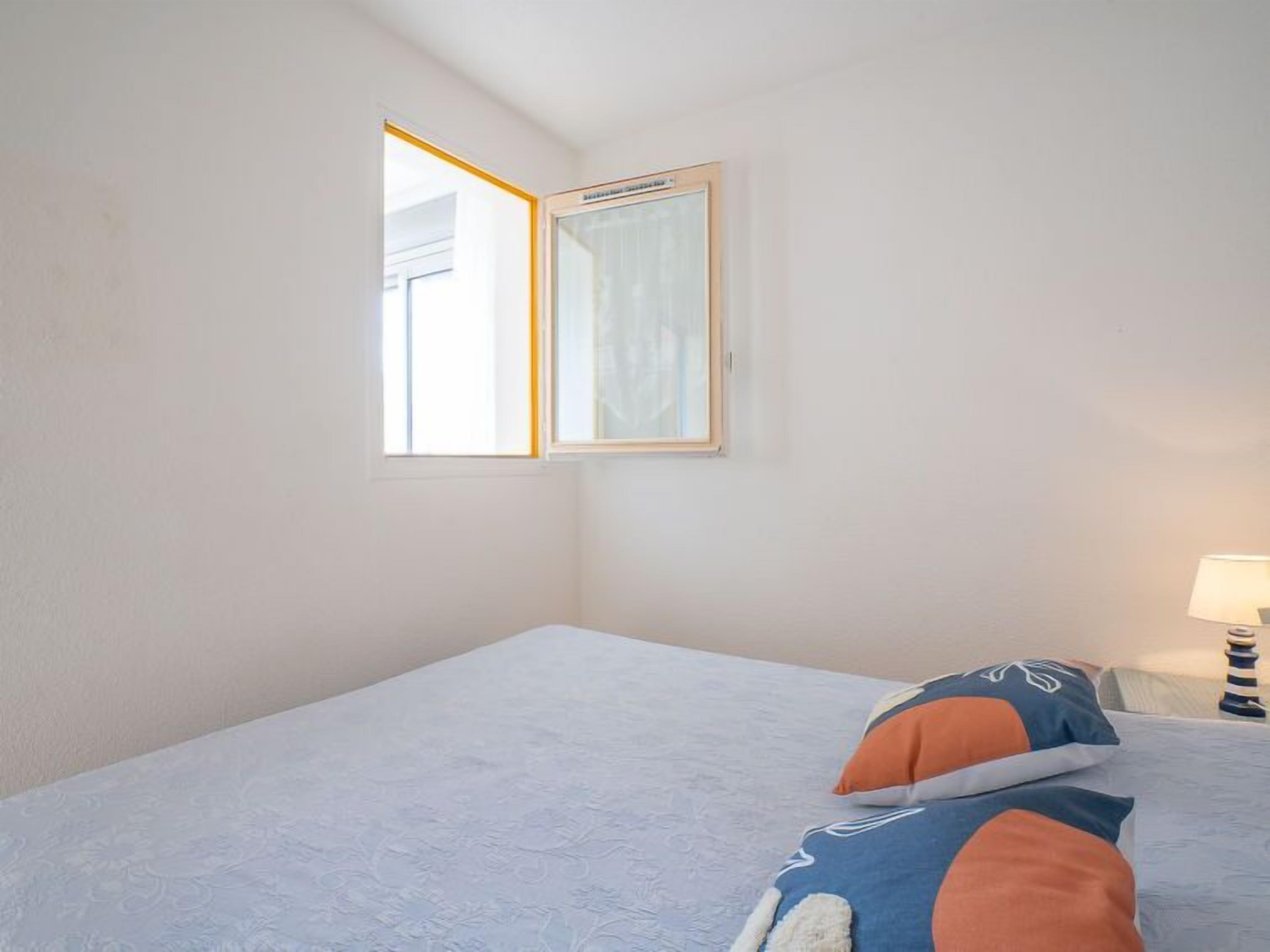Une pièce simple et épurée, avec un lit, une fenêtre et une table de chevet sur laquelle se trouve une lampe.