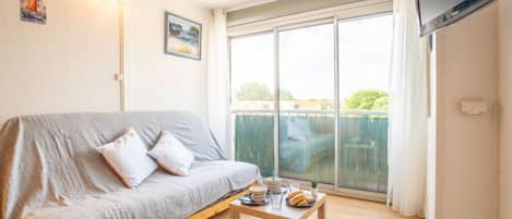 A cozy living room with a grey sofa, a wooden coffee table with snacks, and a large sliding glass door leading to a balcony.