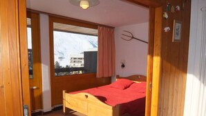 Un lit en bois avec un couvre-lit rouge, dans une pièce dotée d’une grande fenêtre, de murs en bois et offrant une vue sur les bâtiments environnants.