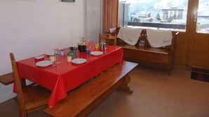 Un coin repas avec une table rouge dressée pour quatre personnes, des bancs en bois, une fenêtre donnant sur un paysage enneigé et un tableau encadré au mur.