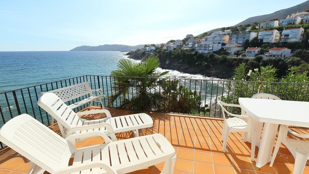 Une terrasse avec des chaises et une table en plastique blanc, donnant sur une zone côtière avec des maisons et une vue sur la mer.