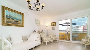 Un vaste salon avec un canapé blanc, des fauteuils à motifs et un lustre. On aperçoit un balcon aménagé avec du mobilier d’extérieur à travers une porte vitrée.