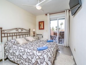Un dormitorio con cama, mesitas de noche, lámpara, ventilador de techo, televisor y balcón con sillas.